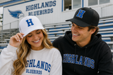 Two smiling young adults standing in front of a school building with bleachers. The woman wears a white sweatshirt and white baseball cap with blue 'HIGHLANDS BLUEBIRDS' text and a large blue 'H' logo.