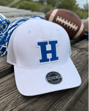 A white trucker-style baseball cap with a large blue embroidered letter H on the front, resting on a wooden picnic table outdoors with a football and blue-and-white tassels nearby.